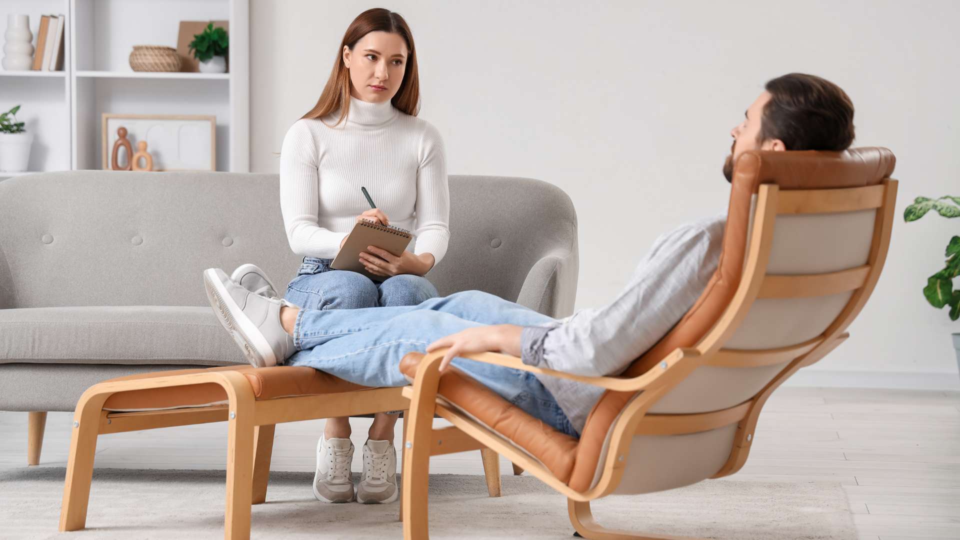 Therapist taking notes in a notebook while client relaxes in a wooden lounge chair during a session in a bright living room.