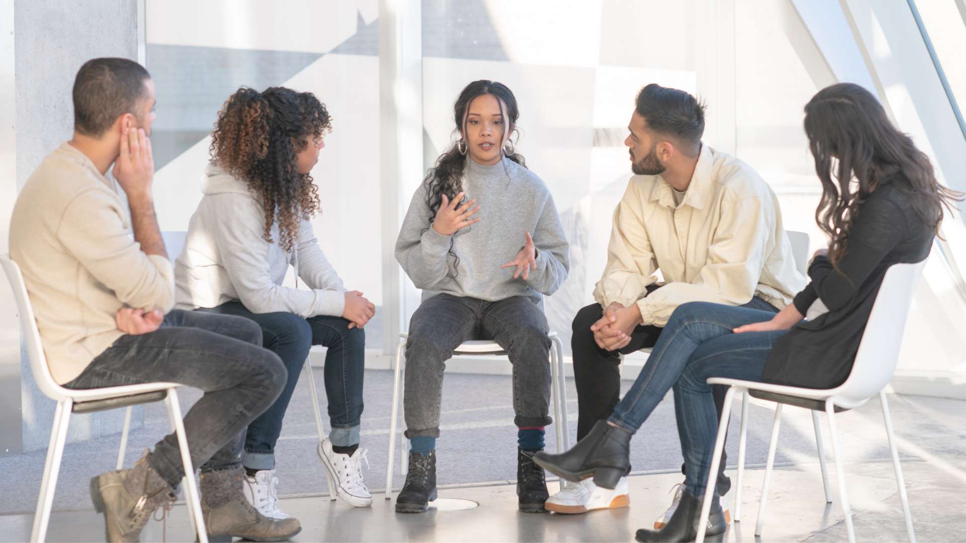 Five people seated in a circle, a woman in a gray sweatshirt speaking with hand gestures.