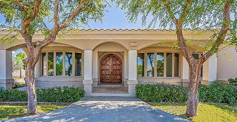 Beige single-story house with an arched central doorway, flanked by windows, manicured hedges, and two trees over a concrete path to the door