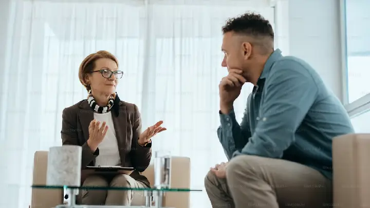 Professional woman wearing glasses explains something to a man in a blue shirt during a meeting in a bright office setting.
