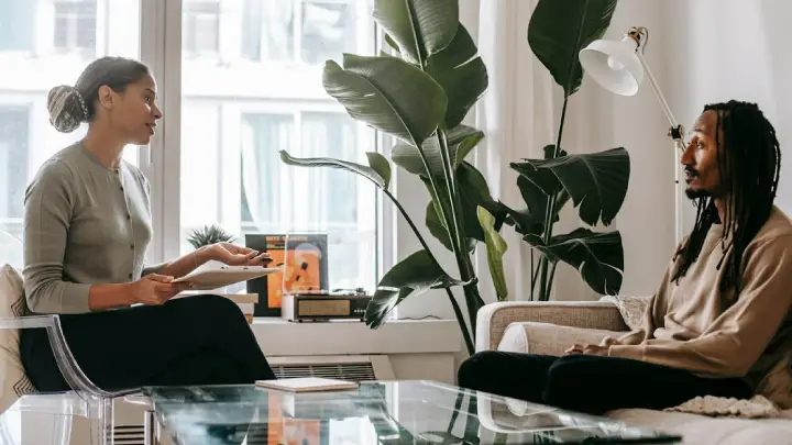 Two people sit across from each other in a bright living room; a woman holds papers while talking to a man with dreadlocks, with large plants behind them.