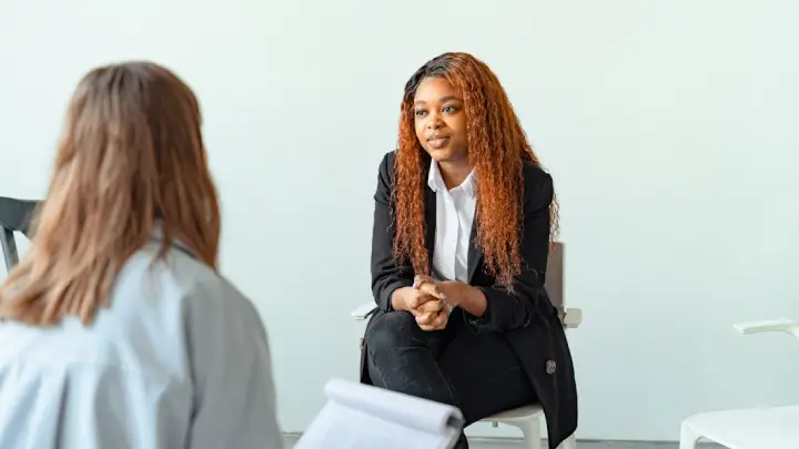Two professionals converse in a bright, minimalist office; a woman with curly hair in a blazer sits facing another person.