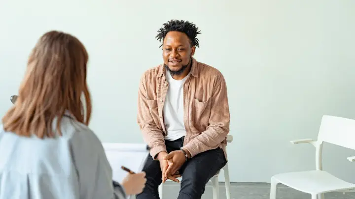 Therapy or counseling session: a man sits on a chair and speaks while a clinician in the foreground takes notes in a bright, minimal room.