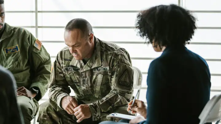 Soldier in camouflage uniform participating in a counseling session with a civilian counselor taking notes in a bright room.