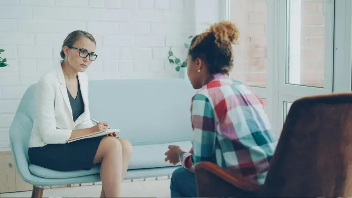 Therapist taking notes during a counseling session with a client seated opposite in a bright room.