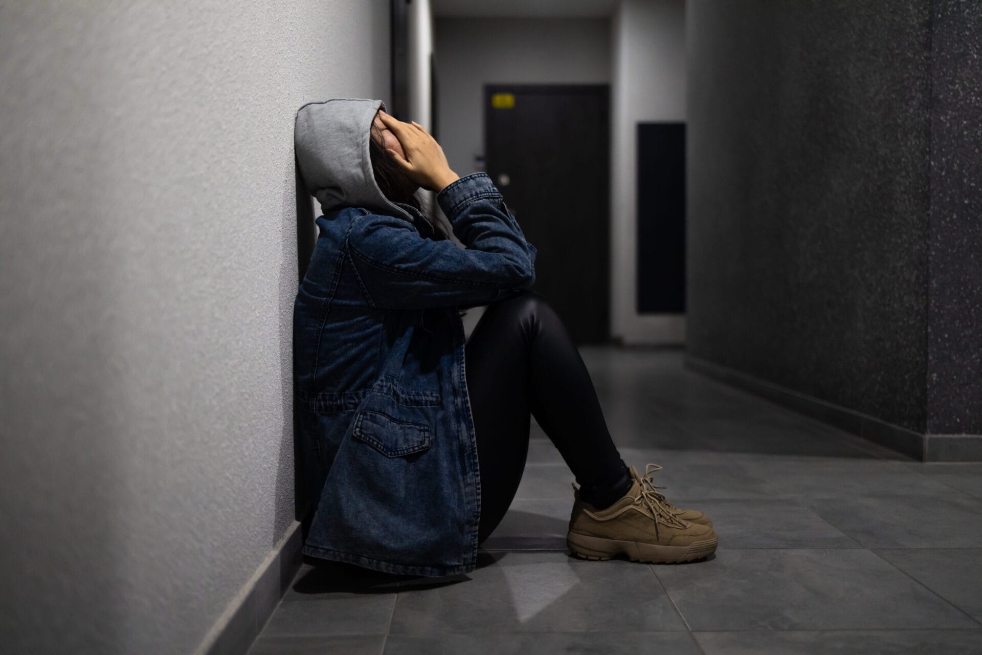 Person sitting on a hallway floor against the wall with a gray hoodie, denim jacket, and hands covering their face, appearing distressed.